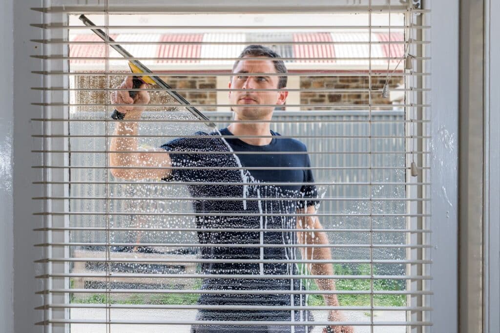 A man using a window cleaner to clean blinds, focusing on window maintenance and using window cleaning tools.